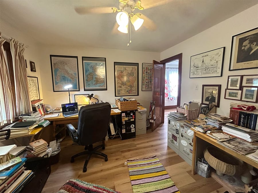A home office with two desks, stacks of books and papers, maps and framed art on the walls, a ceiling fan, and a colorful rug on a wooden floor. A doorway leads to another room with a window and curtain.
