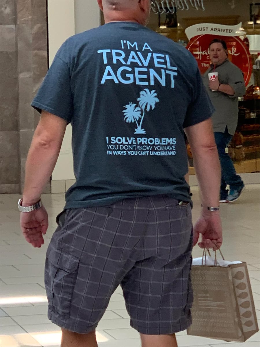 A person wearing a shirt that says “I’m a Travel Agent. I solve problems you don’t know you have in ways you can’t understand” walks in a mall, holding a shopping bag.