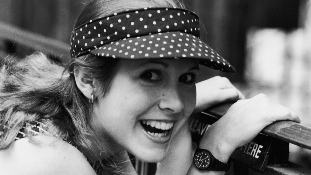 A young woman in a polka dot visor smiles brightly at the camera, leaning on a railing. She wears a sleeveless top and a watch, with her hair tied back. The image is in black and white.