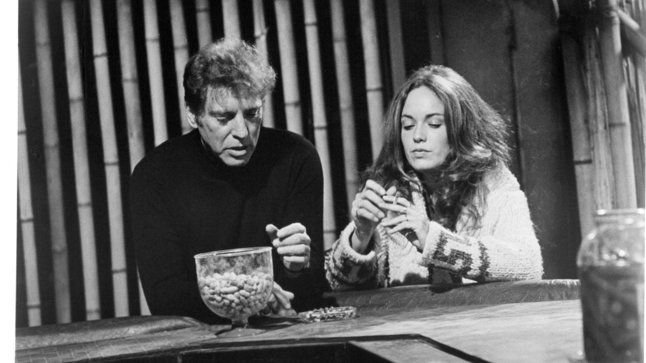 A man and a woman sit at a table; the man is holding a glass container of peanuts, and the woman is cracking open a peanut. Both appear focused on their task in a black-and-white setting.