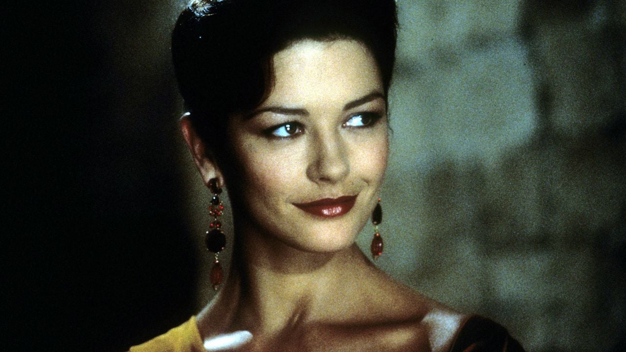 A woman with dark hair in an elegant updo, wearing red and gold drop earrings and bold red lipstick, looks to the side with a slight smile in soft, dim lighting against a blurred stone background.