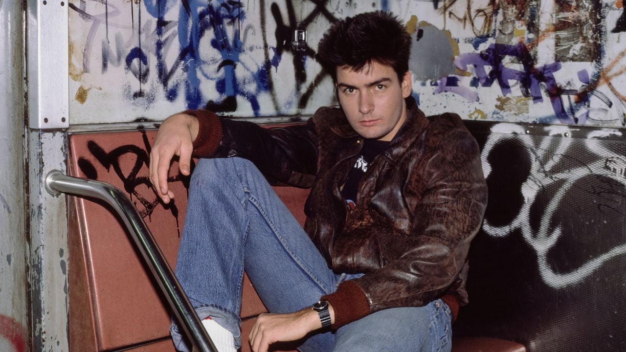 A young man in a brown leather jacket and jeans sits on a subway bench. The wall behind him is covered in colorful graffiti.
