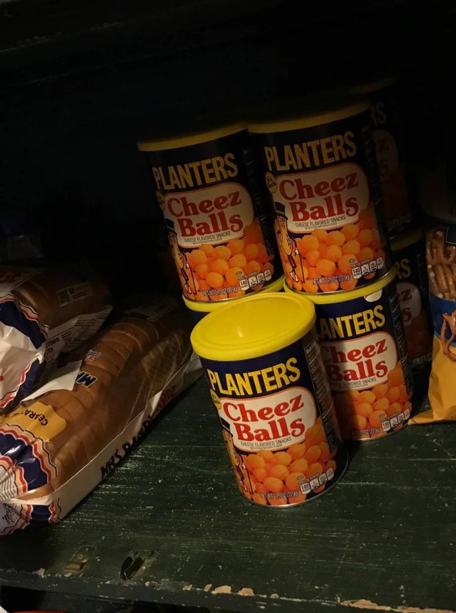 Cans of Planters Cheez Balls are stacked on a dark green shelf next to loaves of bread and a bag of pretzels.