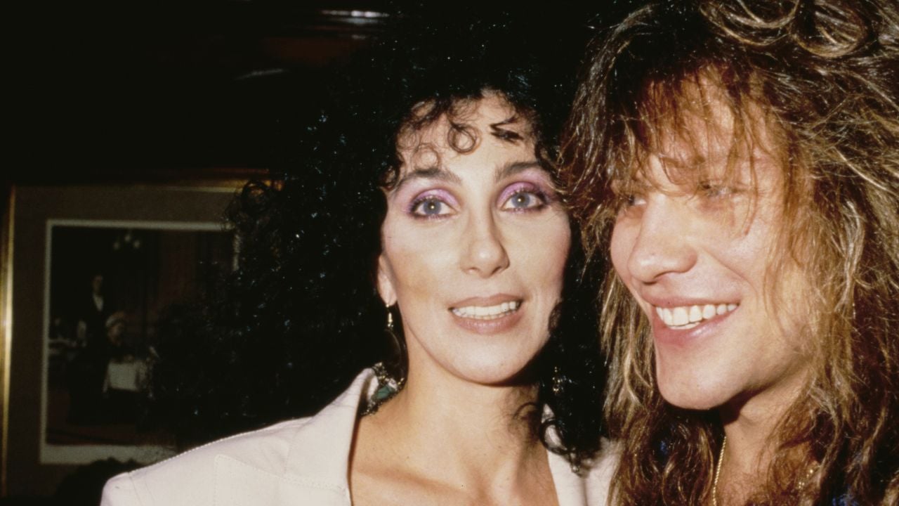 Two people with big, curly hair stand close together, smiling. The person on the left has dark curly hair and purple eye makeup; the person on the right has lighter, wavy hair and is grinning. An old photo is visible in the background.