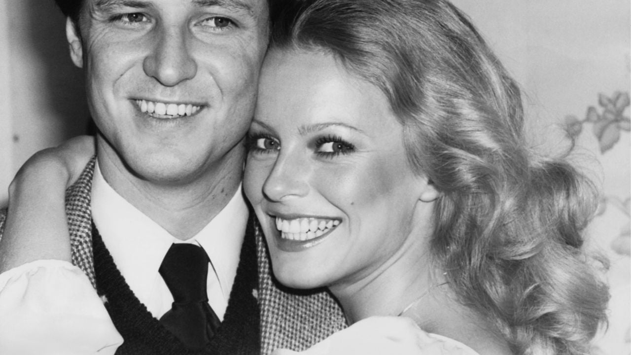 A black-and-white photo of a smiling man in a suit and tie standing next to a woman with long wavy hair, who is also smiling and has her arm around his shoulder.