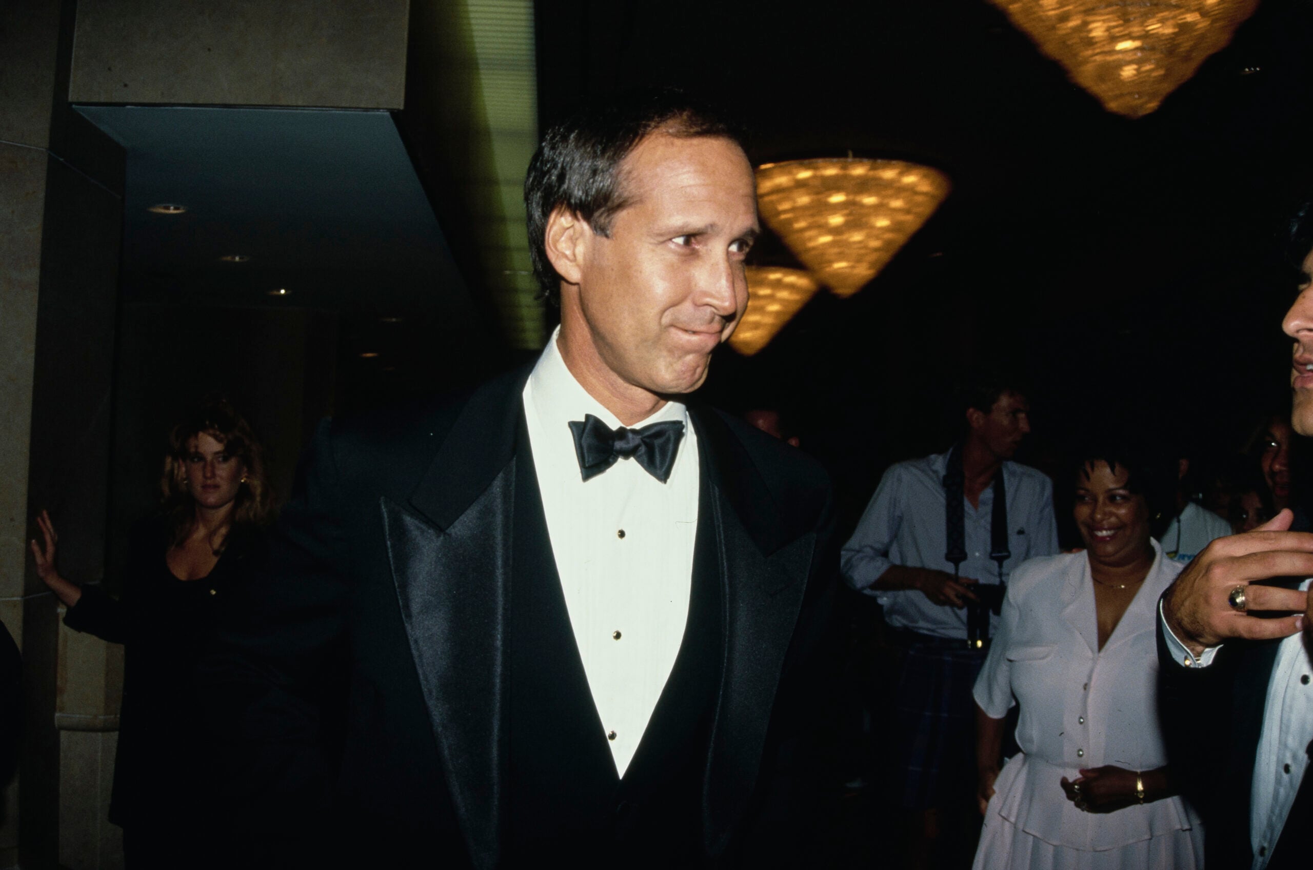 A man in a black tuxedo and bow tie stands indoors at a formal event, surrounded by other people dressed in formal attire. The background features several large, glowing chandeliers.