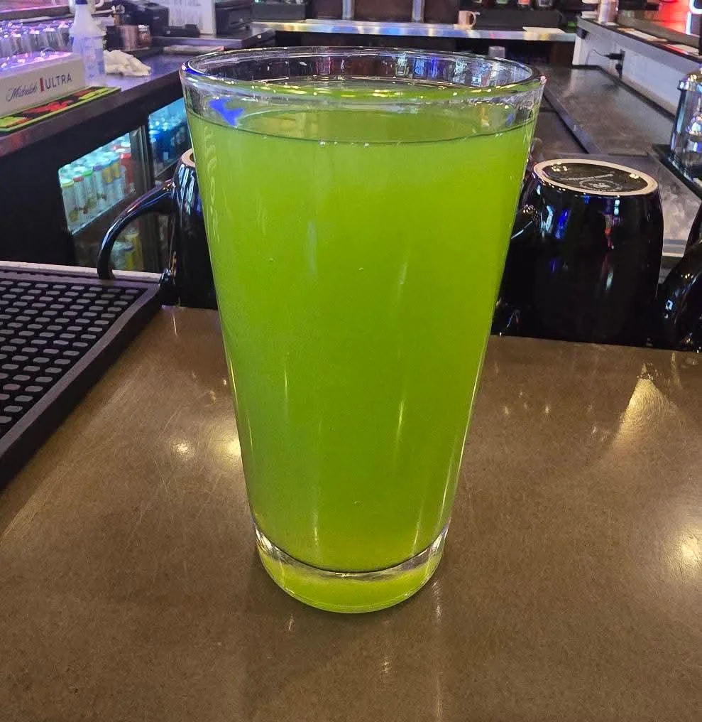 A pint glass filled with a bright green beverage sits on a bar counter, with bar equipment and drinks visible in the background.