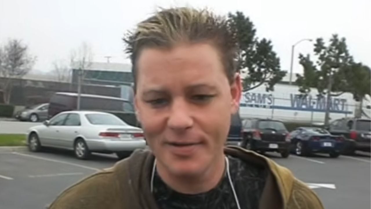 A person with short, spiked hair stands in a parking lot with cars and trees in the background. It appears to be a cloudy day.