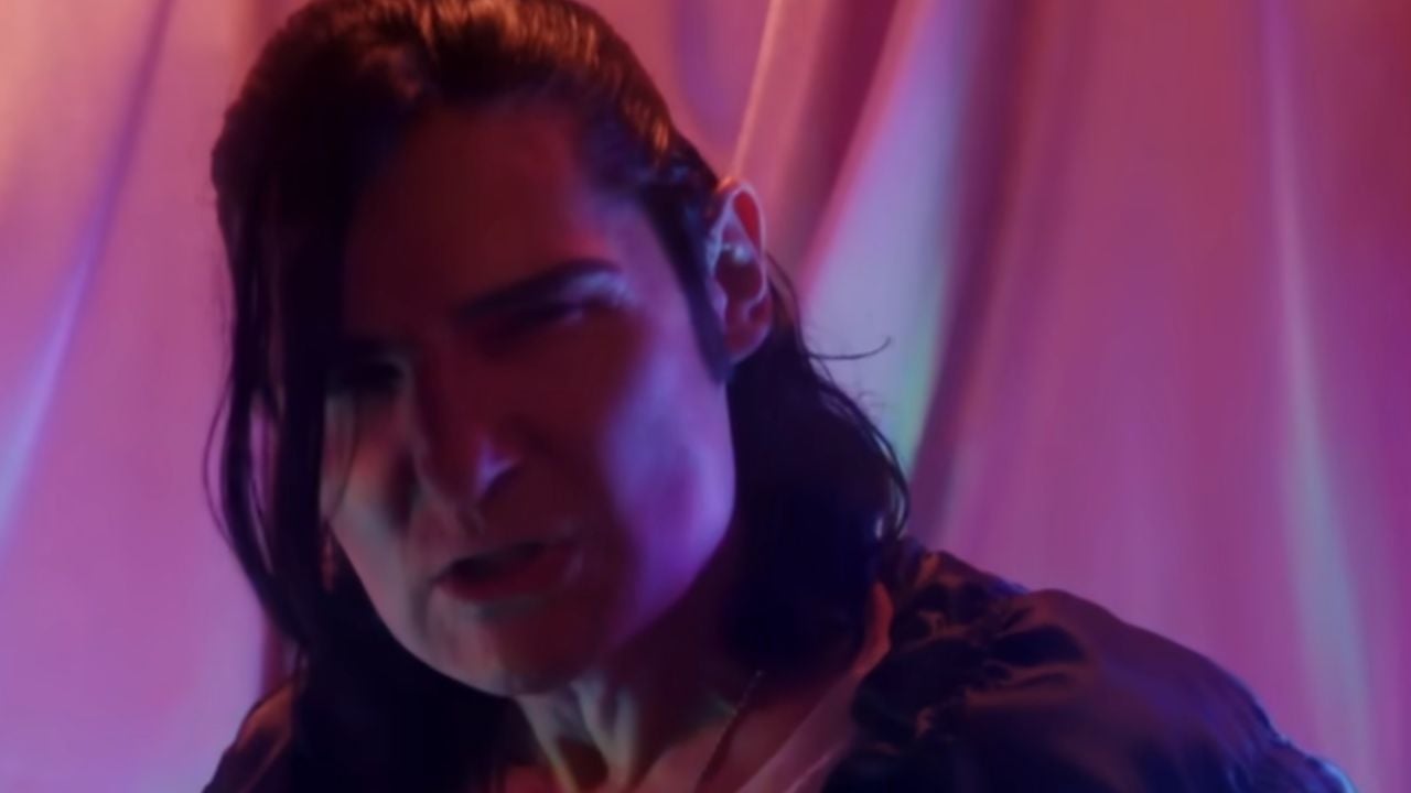 Person with long dark hair and intense expression stands in front of draped fabric, illuminated by pink and purple lighting.