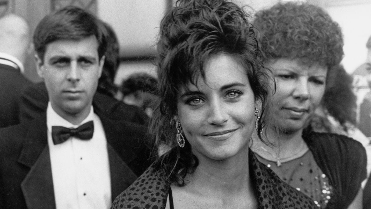 A woman with dark hair and earrings smiles at the camera at a formal event, with two people standing behind her, one in a tuxedo and one in a dress.