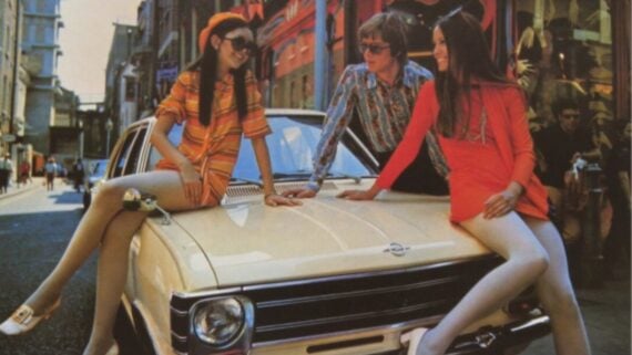 Three people in colorful 1960s-style outfits sit and lean on the hood of a cream-colored classic car parked on a city street, smiling and chatting on a sunny day.