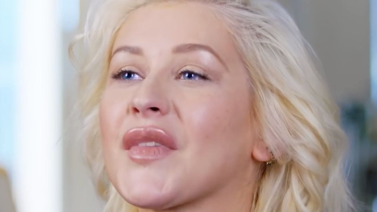 A person with long, wavy blonde hair and blue eyes looks slightly upward. The person has full lips and a natural makeup look, with a soft background behind them.
