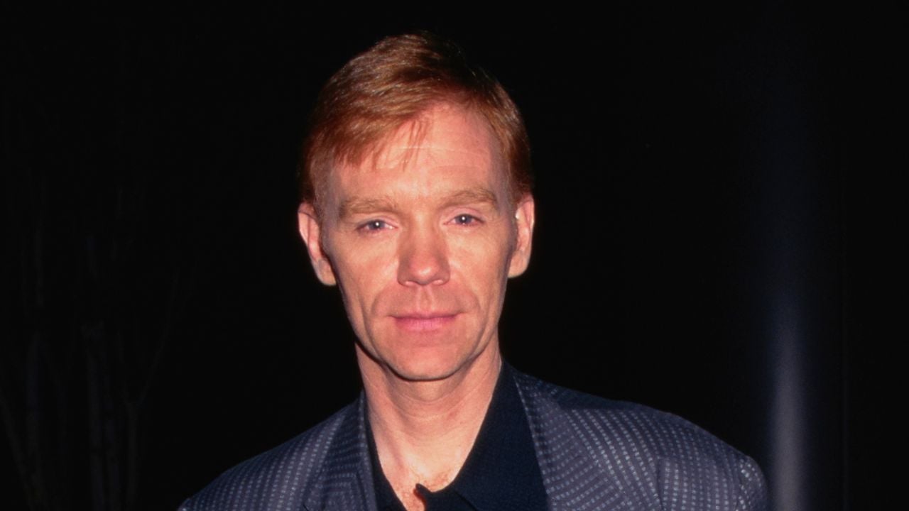 A man with short, red hair wearing a dark suit jacket stands against a dark background, looking directly at the camera with a neutral expression.