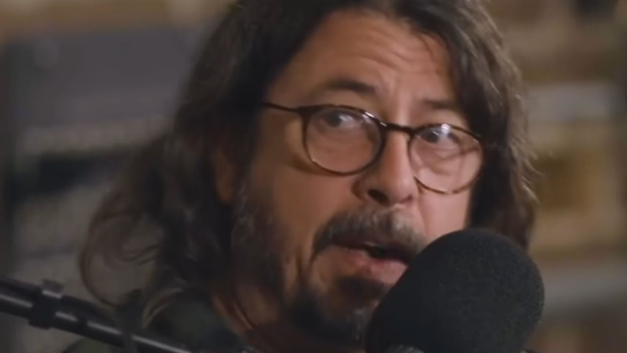 A man with long hair, glasses, and a beard speaks into a black microphone.