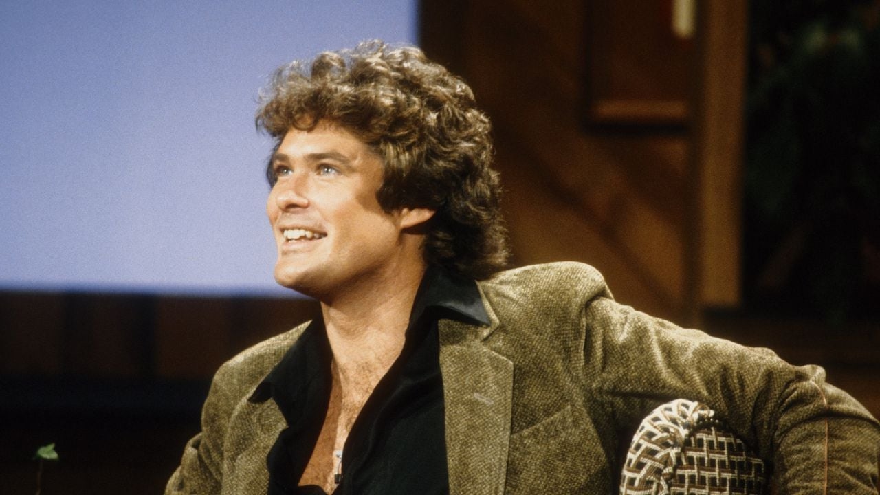 A man with curly brown hair, wearing a brown blazer and black shirt, smiles while sitting in a chair with a wooden background behind him.
