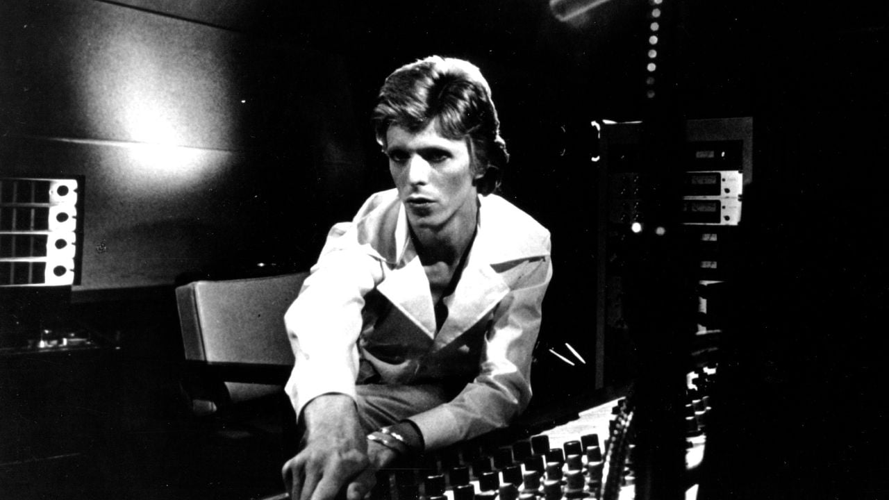 Black and white photo of a person in a suit adjusting controls on a sound mixing board in a recording studio.