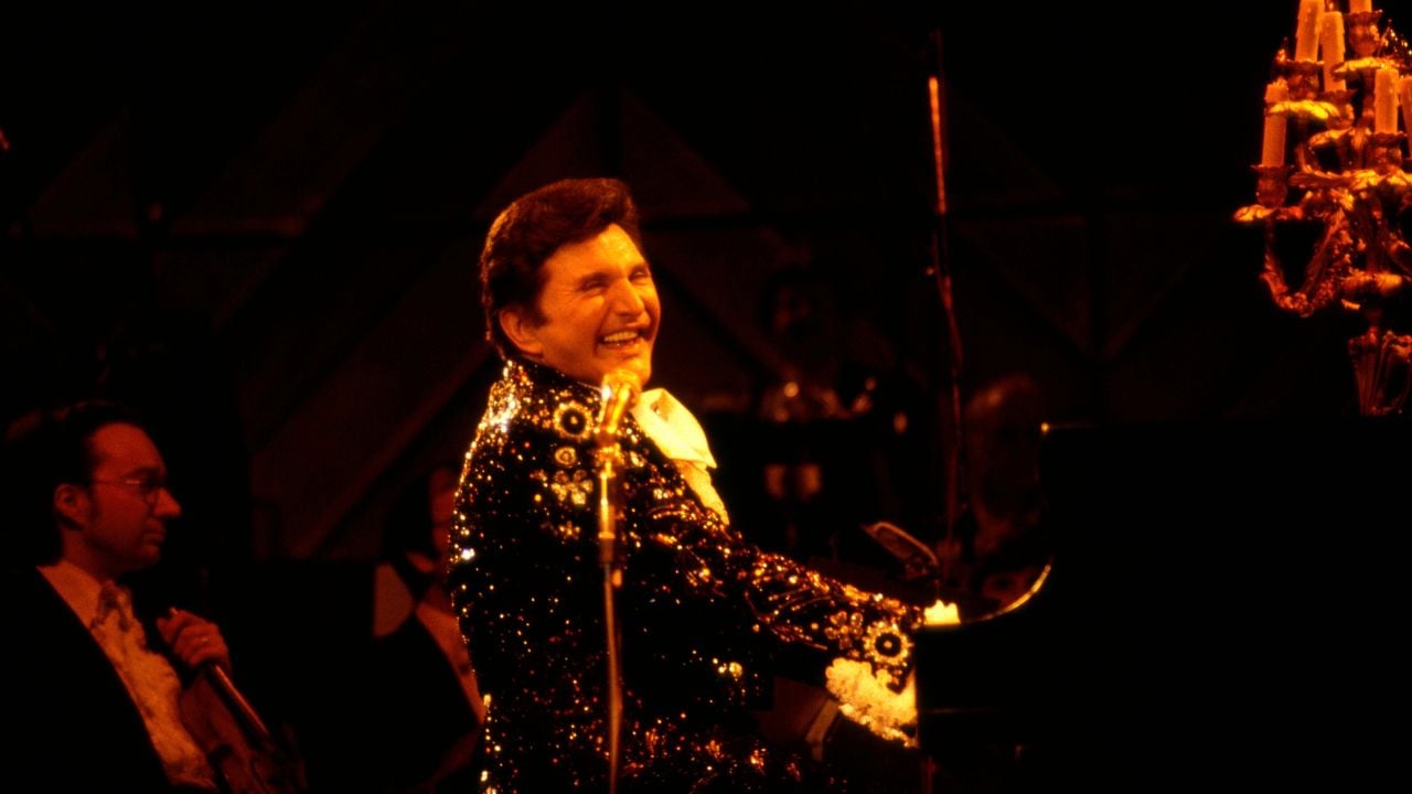 A pianist in a sparkling black jacket smiles while playing a grand piano on a dimly lit stage with a candelabra and musicians in the background.