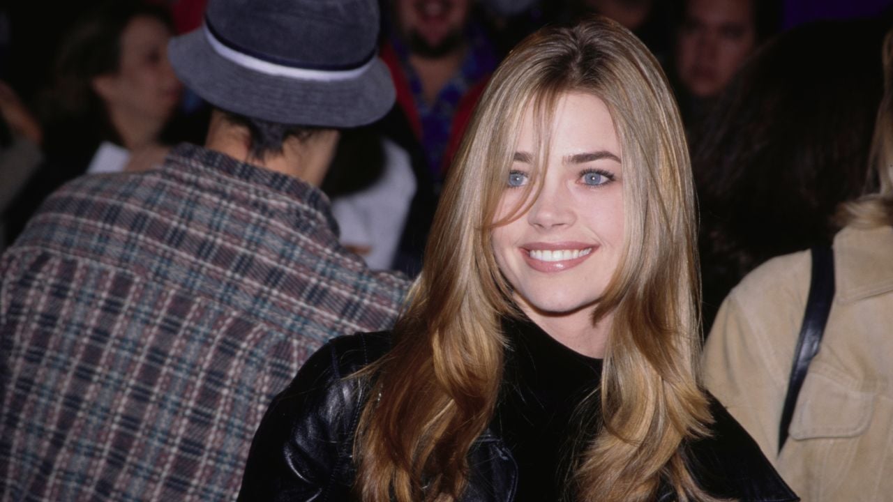 A woman with long blonde hair smiles at the camera, wearing a black leather jacket. She stands in front of a crowd, including a person in a plaid shirt and gray hat facing away from the camera.