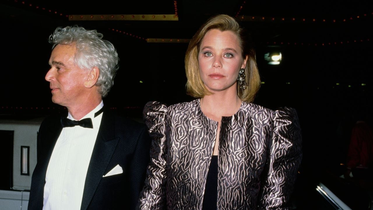 A man in a tuxedo stands next to a woman in a metallic patterned jacket with structured shoulders. Both have serious expressions at a formal event with dim lighting in the background.