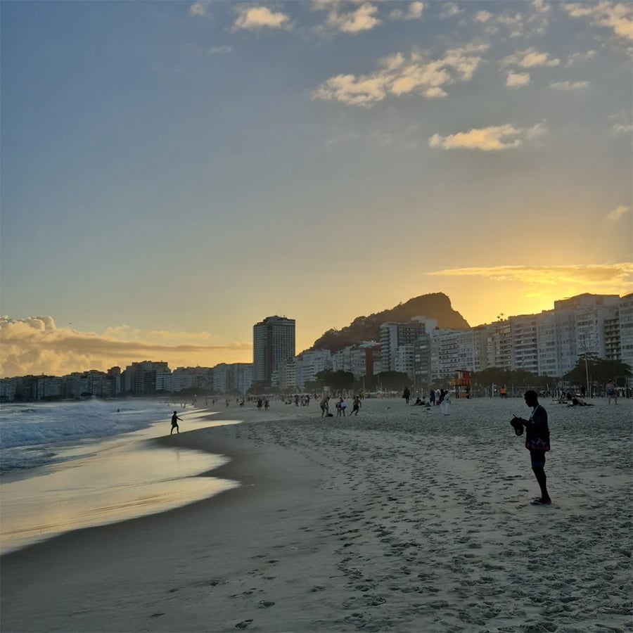 People walk on a sandy beach at sunset, with waves, tall buildings, and a hill in the background under a partly cloudy sky.