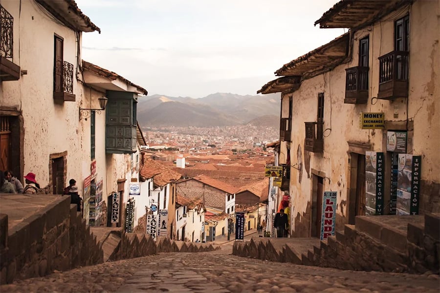 A narrow, cobblestone street lined with old buildings descends toward a city with red rooftops and distant mountains under a cloudy sky. Signs for local businesses hang on the walls.