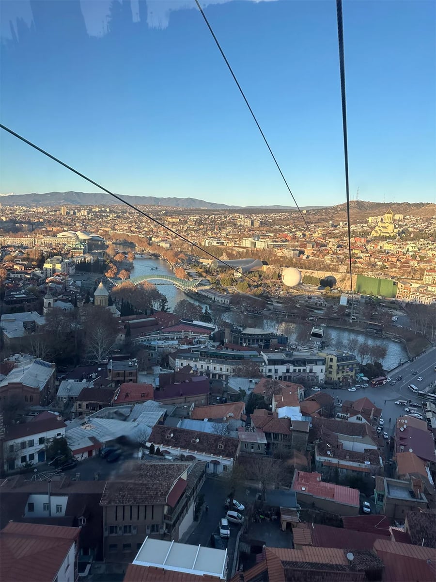 Cityscape view from a cable car, showing red-roofed buildings below, a winding river, roads, and distant mountains under a clear blue sky.