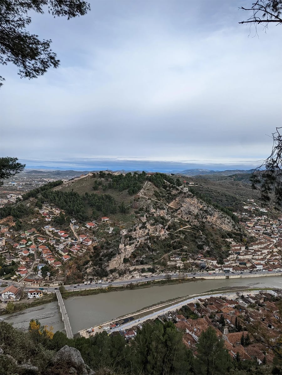 View from above of a town with red-roofed houses, a hill with trees, ancient fortress ruins, and a river with a bridge crossing it under a cloudy sky.