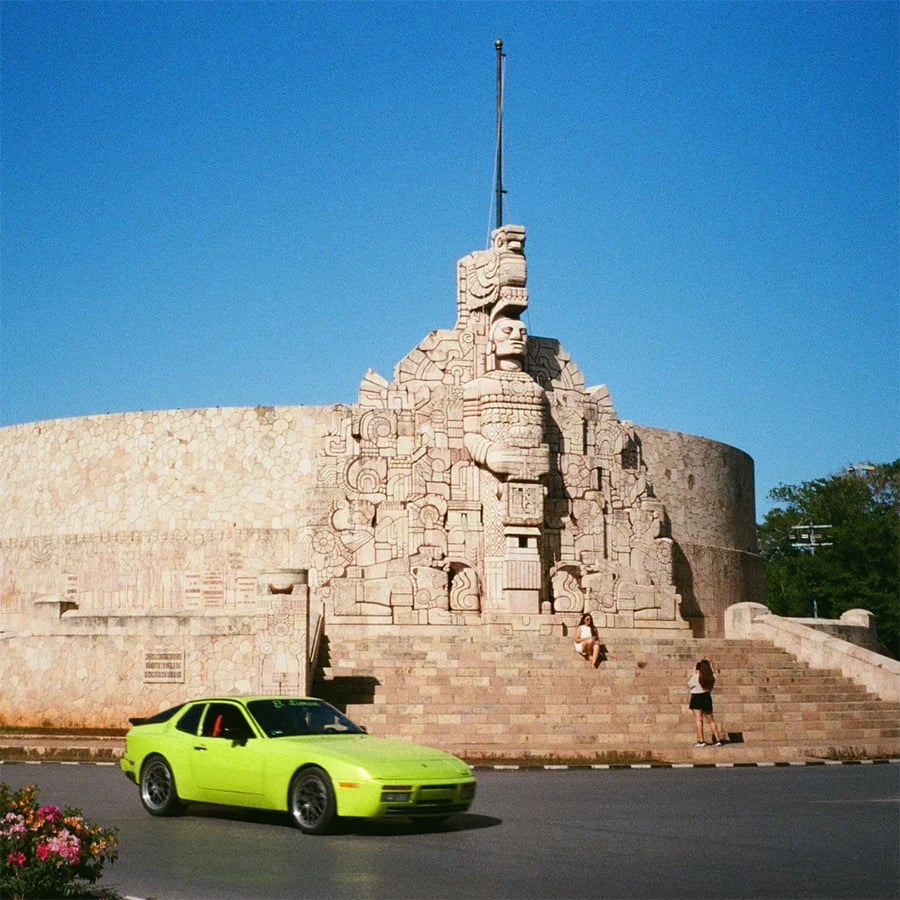A bright green car drives past the Monumento a la Patria, a large stone monument with intricate carvings, under a clear blue sky. Two people sit on the steps in front.