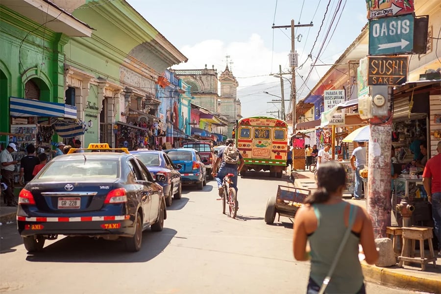 A busy street scene in a colorful town with cars, a bus, people walking, a cyclist, storefronts, and signs including one for sushi. Buildings are painted in green and yellow shades. The sky is clear and sunny.