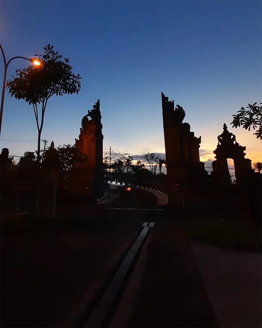 A road curves through a traditional split stone gate at dusk, with a streetlamp glowing and silhouettes of trees and structures against a colorful evening sky.
