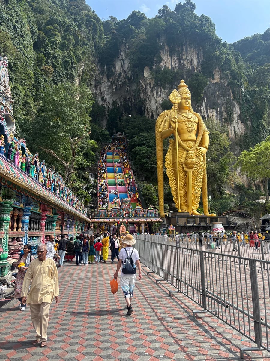Tourists walk toward the colorful staircase and large golden statue of Lord Murugan at Batu Caves in Malaysia, surrounded by lush greenery and temple decorations.