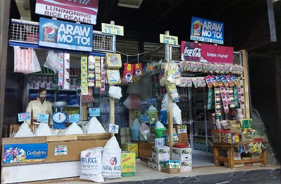 A small Filipino sari-sari store displays bags of rice, hanging snacks, drinks, and household goods. Colorful signs and price tags are visible, with a shopkeeper standing behind the counter.