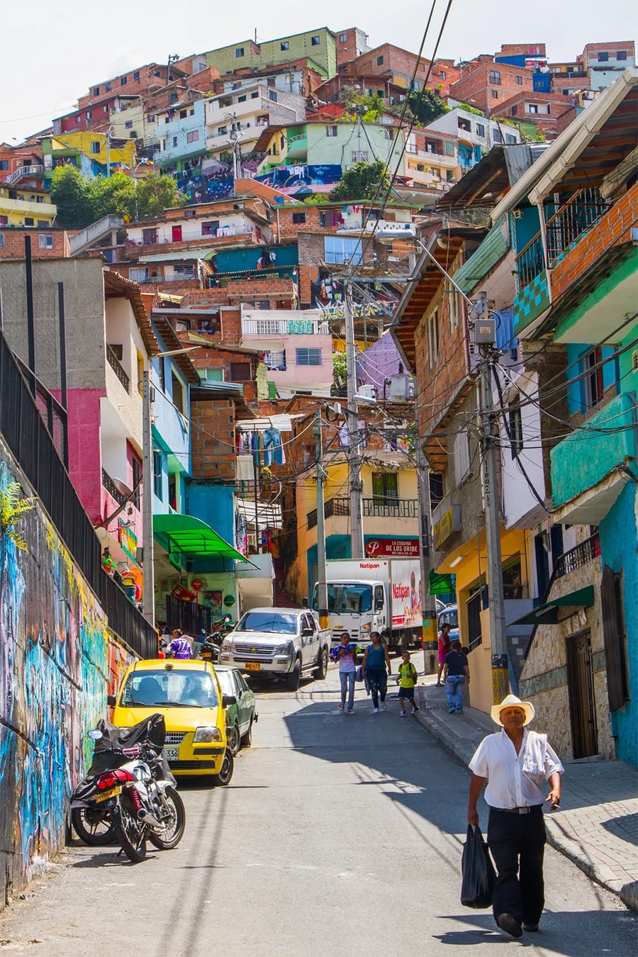 A steep street in a colorful hillside neighborhood with houses stacked closely, cars and motorcycles parked, pedestrians walking, and vibrant murals on the walls.