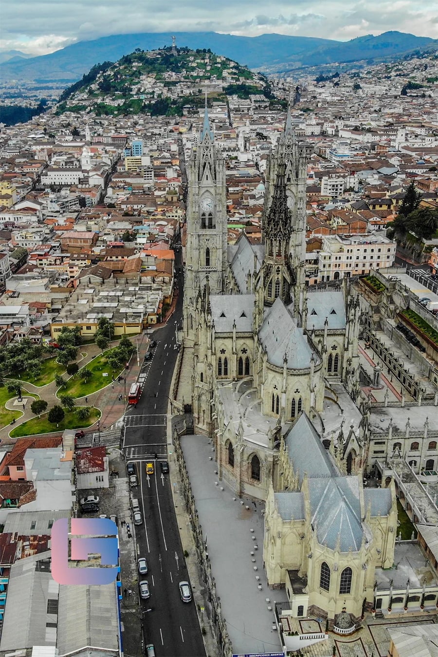 Aerial view of a large gothic-style cathedral with tall spires in a city surrounded by mountains and dense buildings, with roads and cars nearby.