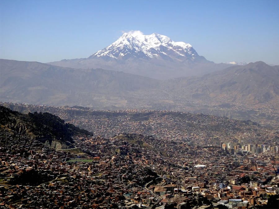 A sprawling cityscape with densely packed buildings sits beneath a towering, snow-capped mountain under a clear blue sky.