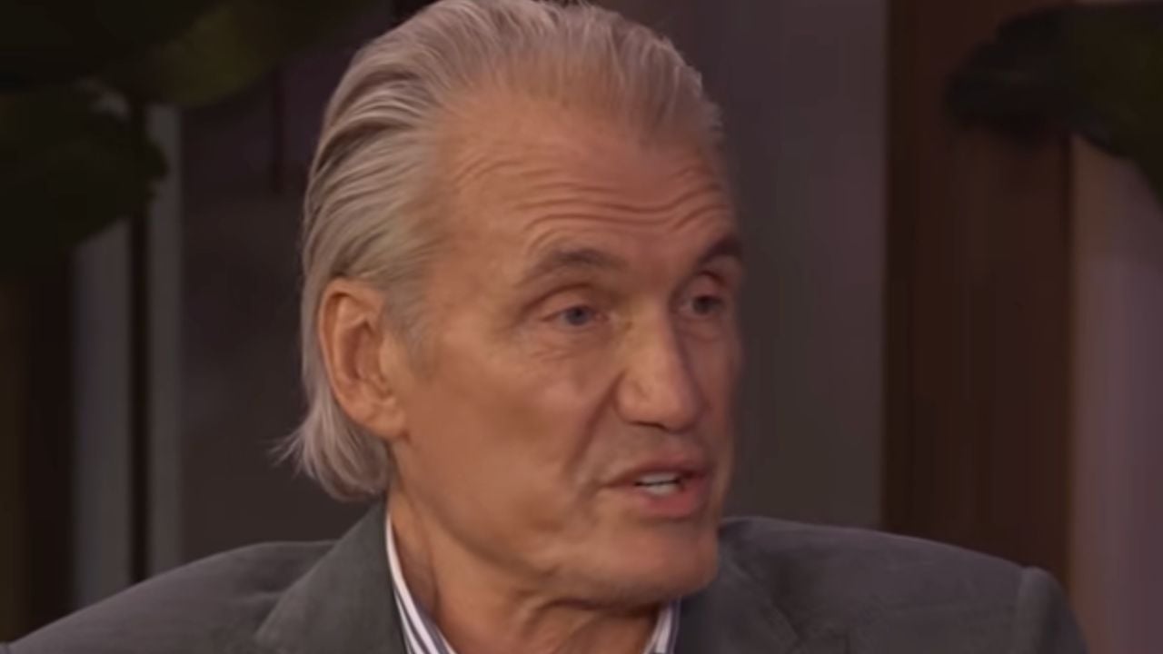 An older man with gray slicked-back hair is speaking. He is wearing a dark blazer and a striped shirt, and is pictured in a softly lit indoor setting.