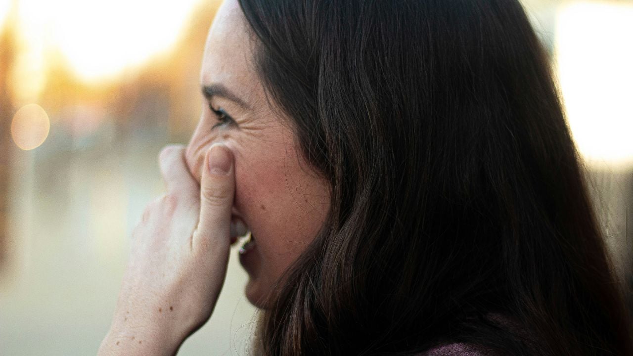 A woman with long brown hair smiles and covers part of her face with her hand. She is outdoors and appears to be laughing, with a soft, blurred background.