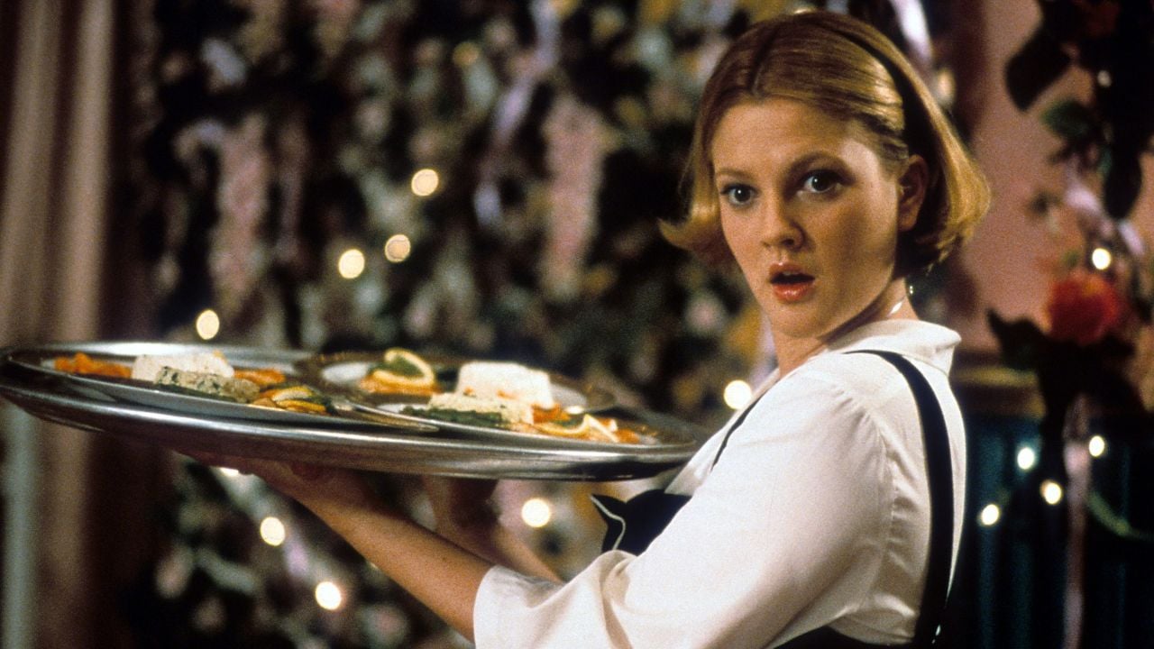 A young woman with short blonde hair, wearing a white shirt and black vest, holds a large tray with food. She looks surprised, with a blurred, festive background of lights and flowers behind her.