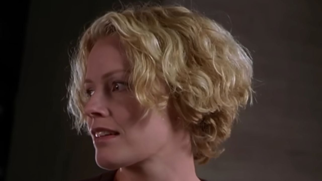 A person with short, curly blonde hair looks to the left, with a neutral expression, against a plain, dark background.