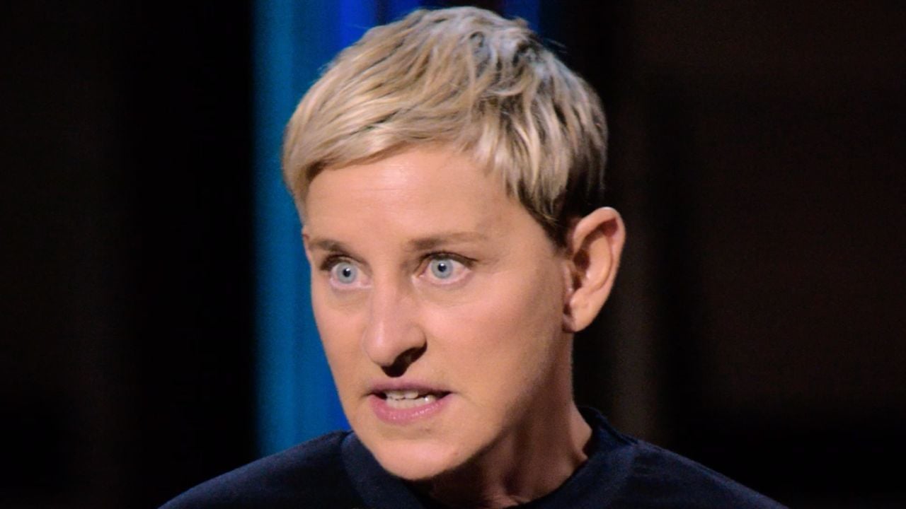 A person with short blonde hair and blue eyes is speaking intensely, looking slightly to the side. The background is dark with a blurred blue vertical line.