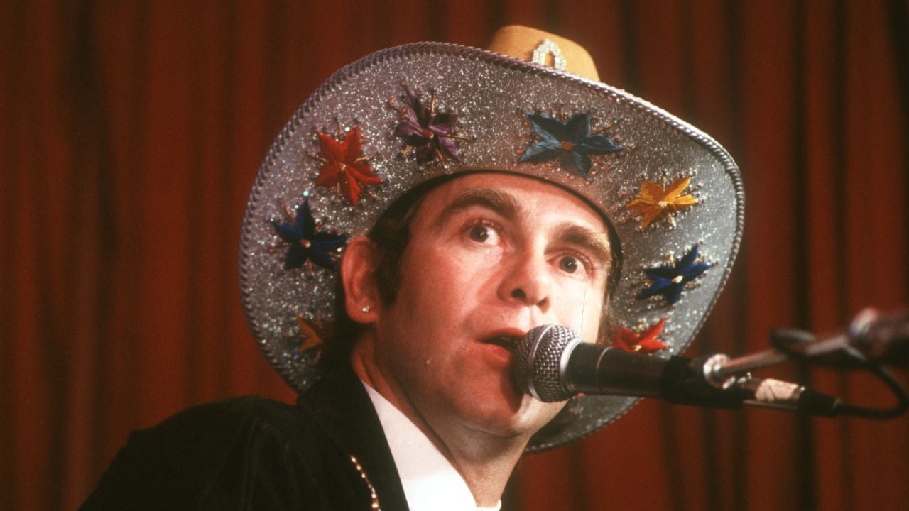 A person wearing a sparkly cowboy hat decorated with colorful stars is singing or speaking into a microphone on stage with a red curtain background.