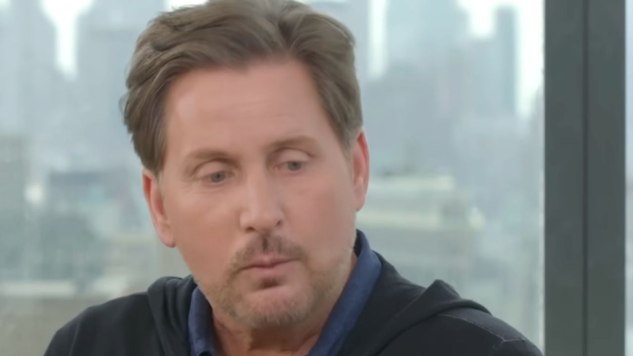 A man with short brown hair and a trimmed beard looks to the side in a brightly lit room with large windows and a blurred cityscape in the background.