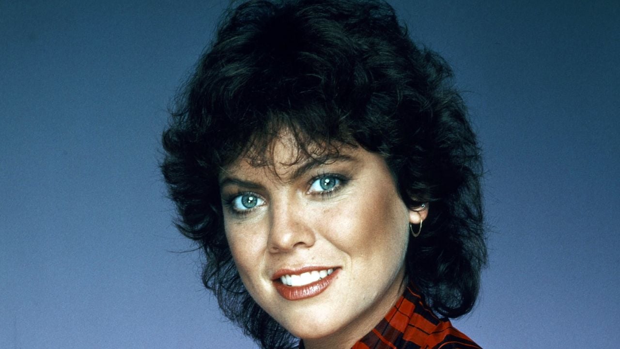 A woman with short, curly dark hair and blue eyes smiles at the camera. She is wearing a red plaid shirt and small hoop earrings, with a blue gradient background behind her.