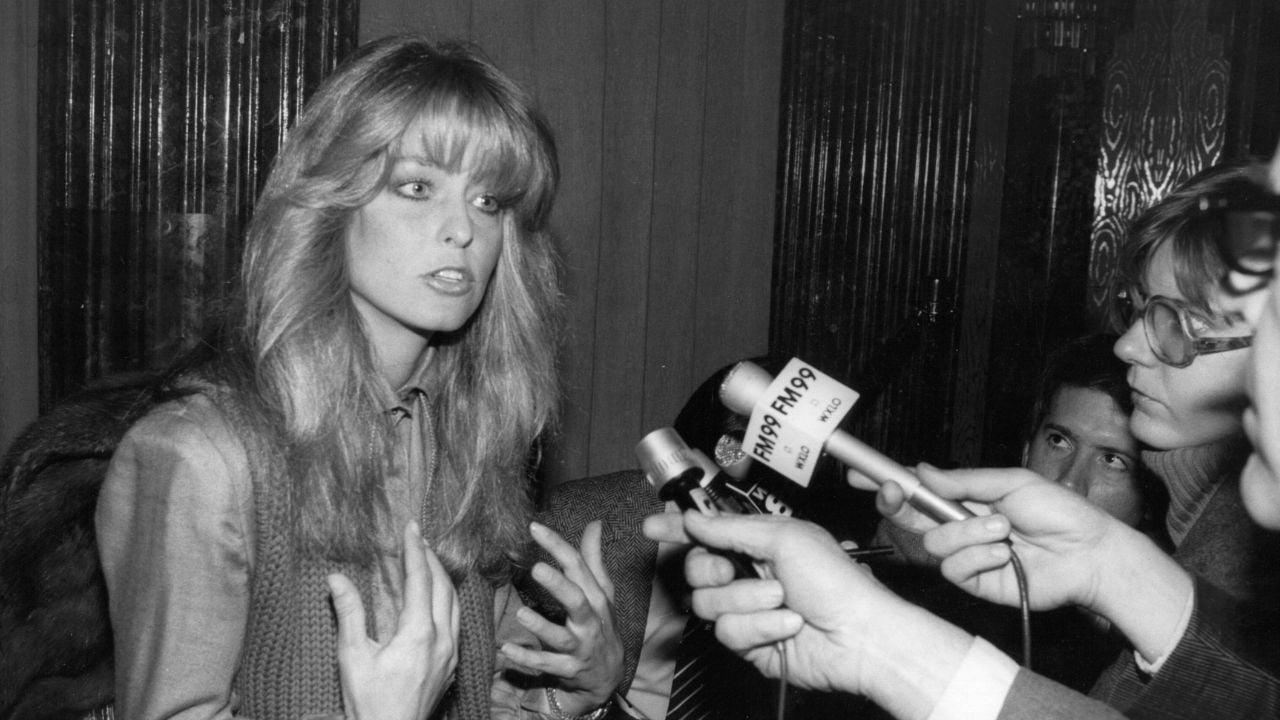 A woman with long, wavy hair speaks to journalists holding microphones during an interview. She gestures with her hands, and several people surround her, holding recording devices and notepads. The photo is in black and white.