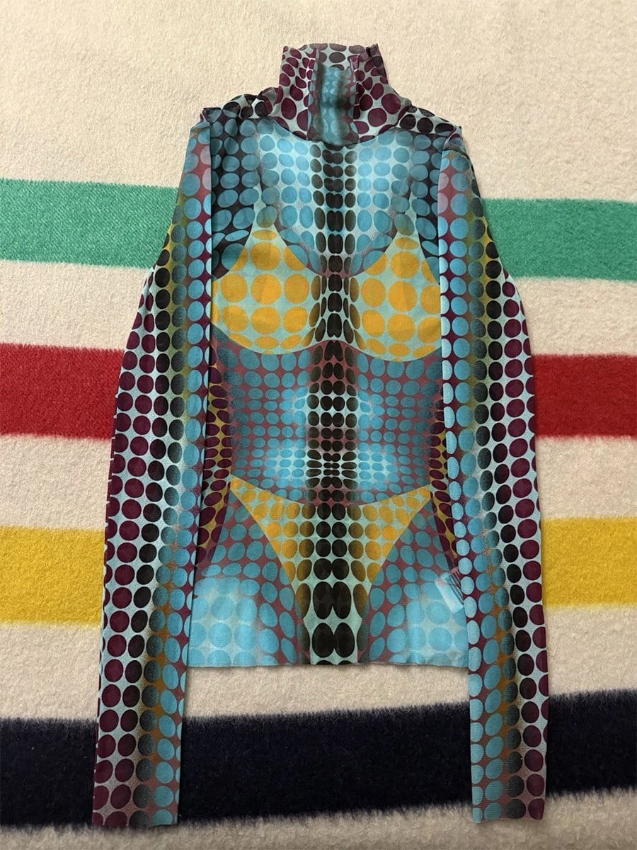 A long-sleeve turtleneck top with a colorful dot pattern that creates an optical illusion of a bikini-clad torso, laid flat on a striped blanket.