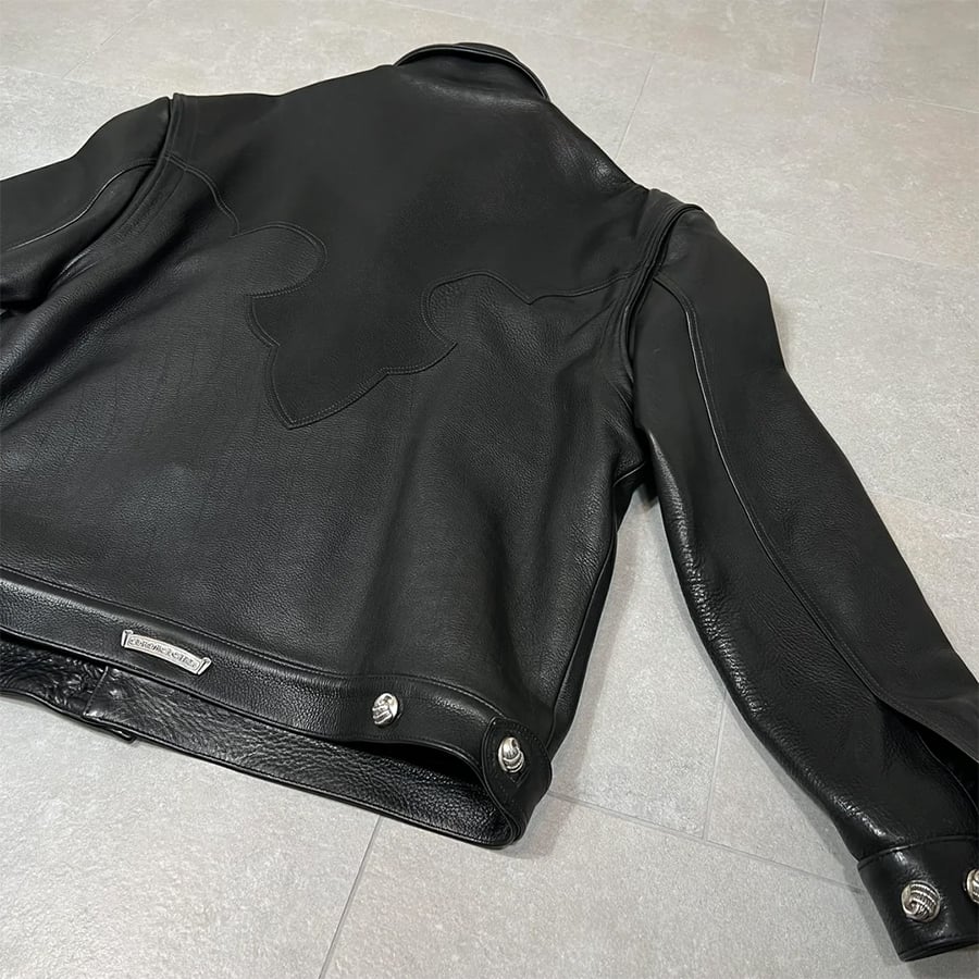 A black leather jacket lies flat on a light gray floor, showcasing its back with a subtle stitched design and metallic button details near the waistband.