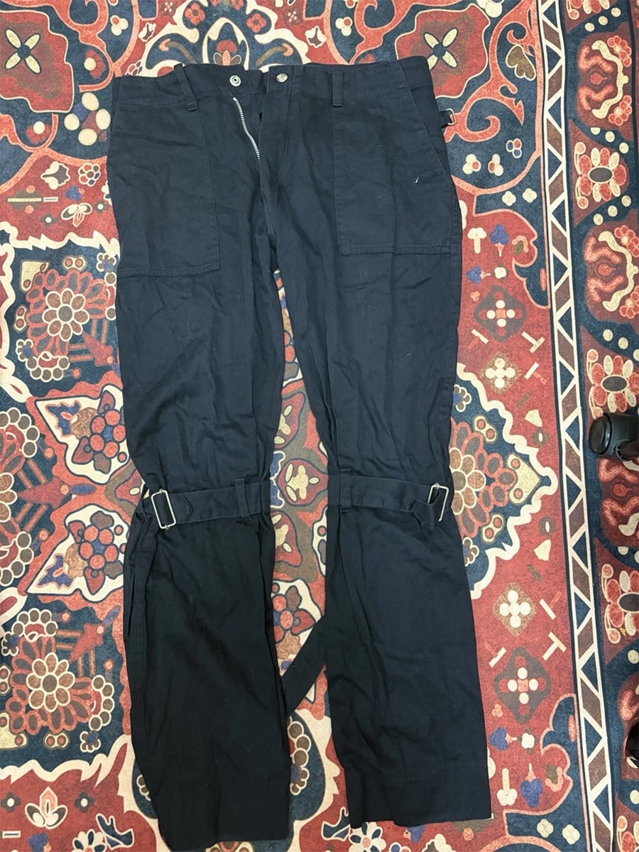 Black cargo-style pants laid flat on a red and blue patterned rug, featuring extra fabric straps and buckles around the knees and lower legs.
