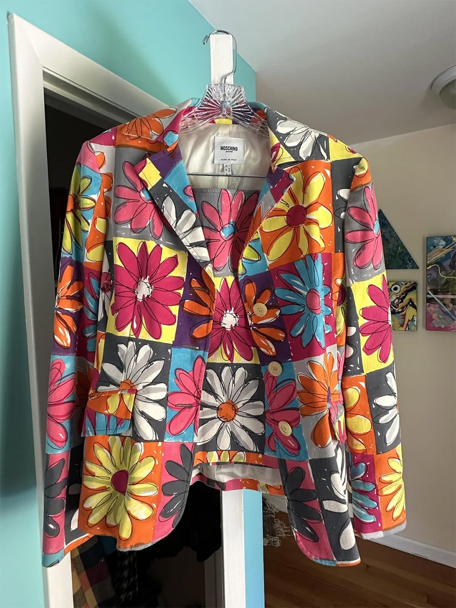 A vibrant jacket with a large, colorful floral pattern in orange, pink, yellow, and white, hanging on a hanger in a room with turquoise walls and artwork on the walls.