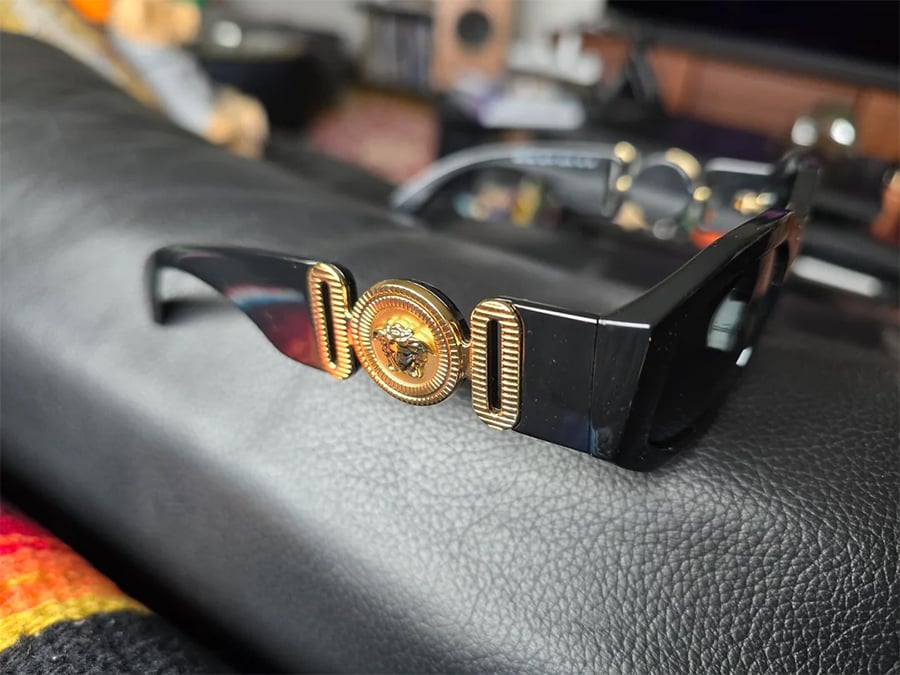 Black sunglasses with thick arms, featuring a gold decorative emblem on the side. The glasses are resting on a textured black surface, with blurred background items visible.