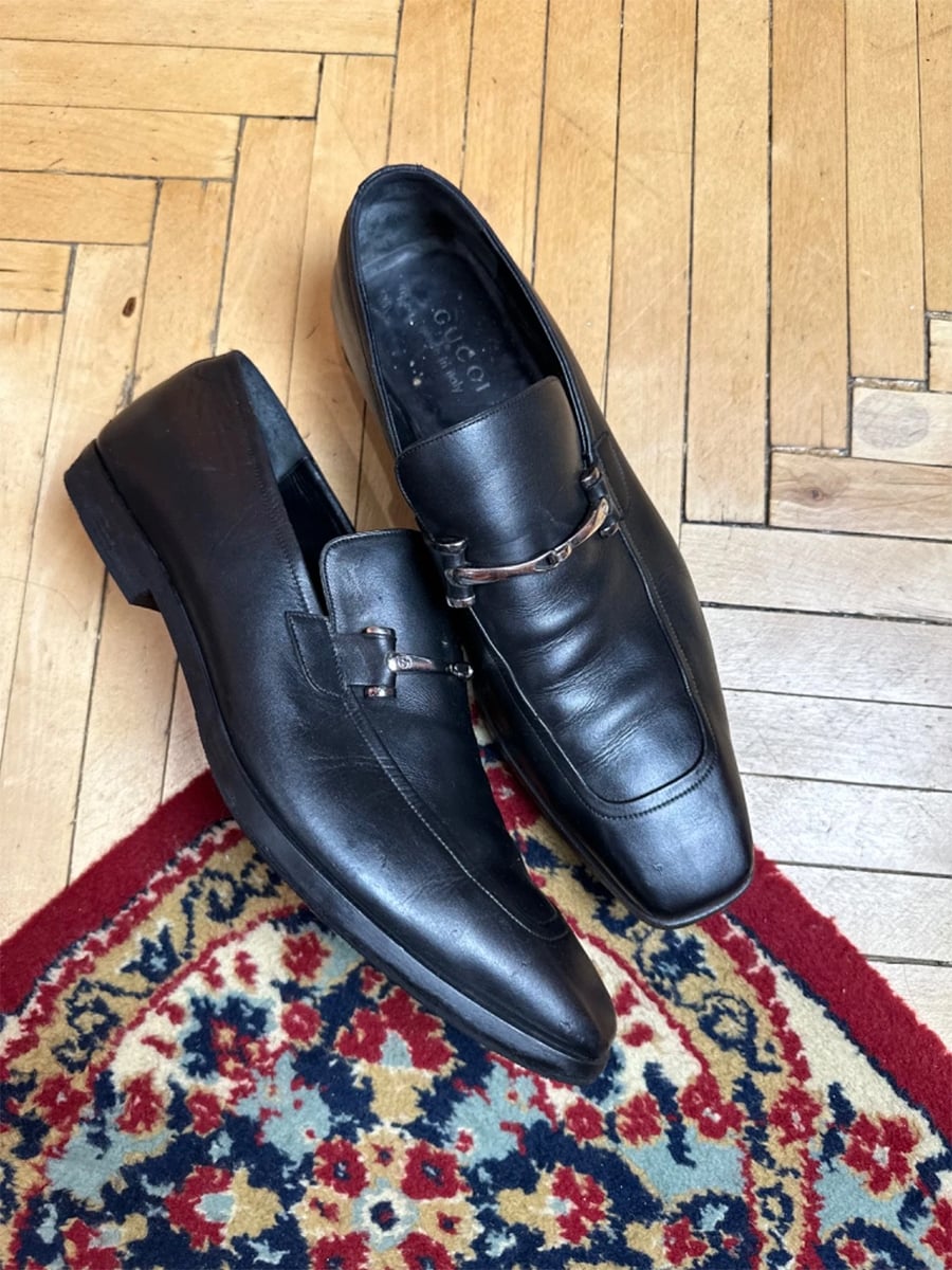 A pair of black leather loafers with metal detailing rests on a wooden floor next to a colorful patterned rug.